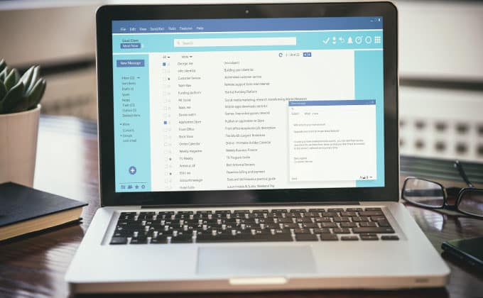A laptop displays an open email inbox and a composed message, with a notebook and plant beside it on the desk