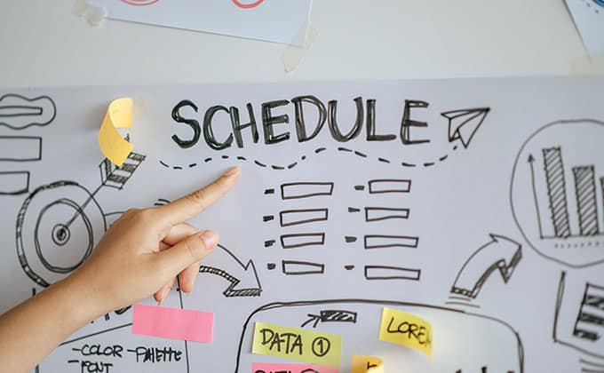 A hand points to a section labeled "SCHEDULE" on a paper with charts, boxes, graphs, and colorful sticky notes.