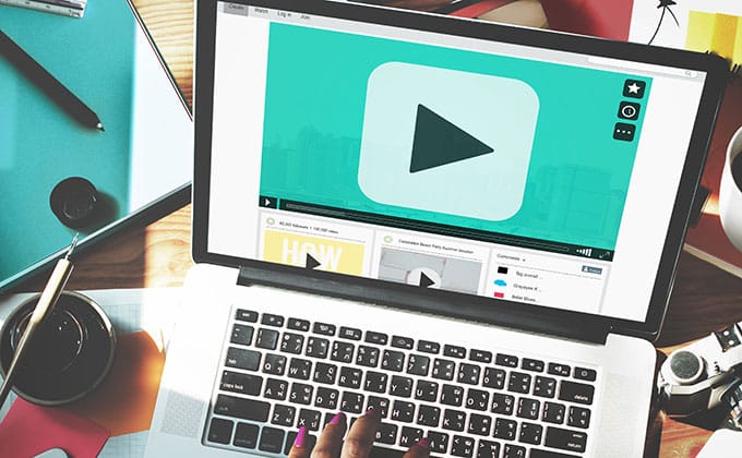 A person uses a laptop displaying an explainer video player screen with a large play button, surrounded by office supplies and paperwork.