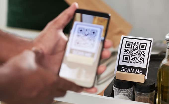 A person engages with QR codes by scanning a "SCAN ME" label on their smartphone at a countertop surrounded by spices and bottles.