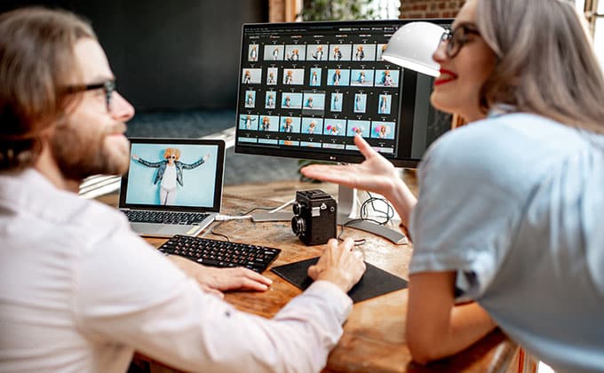 Two people discuss photo selections displayed on a desktop monitor, aiming for higher engagement on their website, with a camera and laptop resting on the wooden desk.