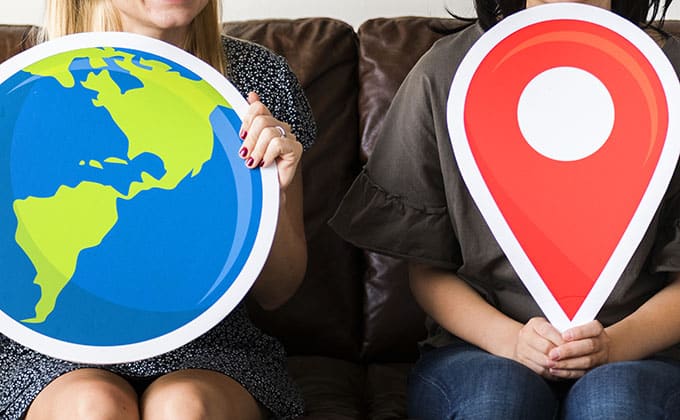 Two people sitting on a couch hold large paper cutouts: one of a globe showing the Americas, the other of a red map location pin, symbolizing how local businesses can reach nearby customers through geotargeted ads.