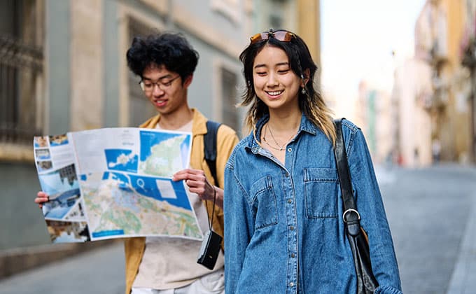 Two young adults walk down a city street past local businesses; one holds a map while the other smiles, both in casual clothes.