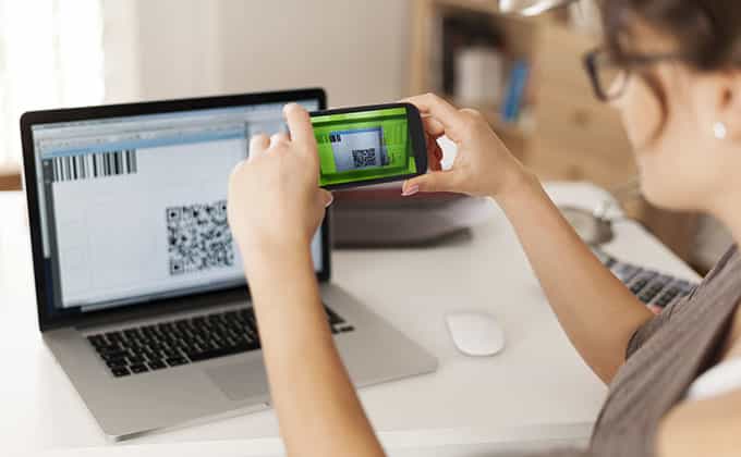 A person uses a smartphone to scan a QR code displayed on a laptop screen at a desk with a computer mouse and papers.