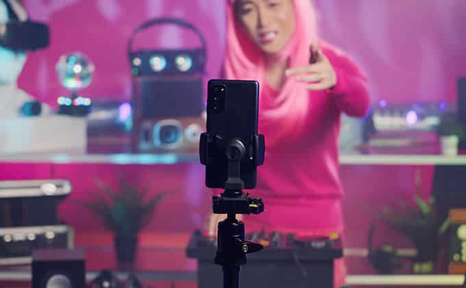 Person with pink hair and a pink sweater livestreaming in a room filled with electronic equipment, filming themselves on a smartphone mounted on a tripod, creating content for Reels or TikTok to boost their business presence.