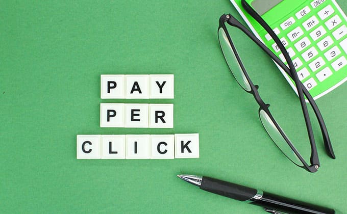 Letter tiles spell “Pay Per Click” on a green background, next to a pair of glasses, a green calculator, and a black pen—perfect for illustrating lead generation or Pay Per Lead Marketing in your complete guide.