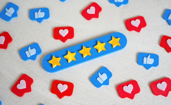 A row of five gold stars on a blue base, surrounded by red heart icons and blue thumbs-up icons, evokes the look of Facebook Reviews and online reviews on a light surface.